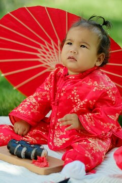 Asian baby in traditional red costume with umbrella, sushi rolls and lantern on picnic blanket