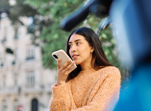 A young woman sits by a parked bicycle in an urban park, holding a smartphone and gazing thoughtfully into the distance. She wears a cozy sweater and jeans, suggesting a relaxed autumn outing. The can
