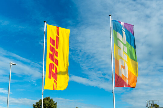 Germany - Aug 22, 2026: DHL flag and a rainbow striped banner waving on tall poles against a blue sky with thin cirrus clouds above a logistics depot at the edge of the frame