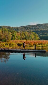 Traveler walking on a dock at Sherman Reservoir Boat Launch in Vermont, USA, surrounded by calm water, autumn hills, reflections, and scenic outdoor landscape. Scenic drone view. Vertical.