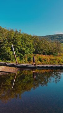Traveler walking on a dock at Sherman Reservoir Boat Launch in Vermont, USA, surrounded by calm water, autumn hills, reflections, and scenic outdoor landscape. Scenic drone view. 4K. Vertical.