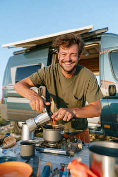 A cheerful man prepares fresh coffee on a portable camping stove outside his parked camper van
