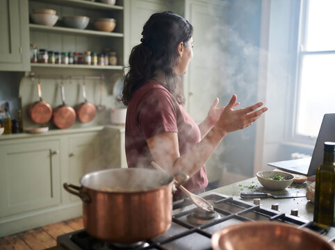 Woman cooking in a home kitchen beside a laptop while steam rises from a large pot, suggesting online learning, remote instruction, or digital recipe guidance.