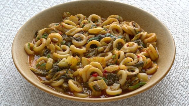 Authentic Italian Anelli Siciliani con Bietole e Pomodori dish. A brown ceramic plate with ring-shaped pasta, slowly rotating on a bright background with a beige geometric pattern.