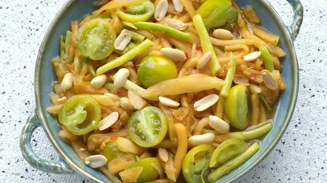 Top view of an alternative Thai som tam salad with stripped orange melon, yellow string beans, green grape cherry tomatoes, and roasted peanuts in a blue-green ceramic bowl. Table spin.