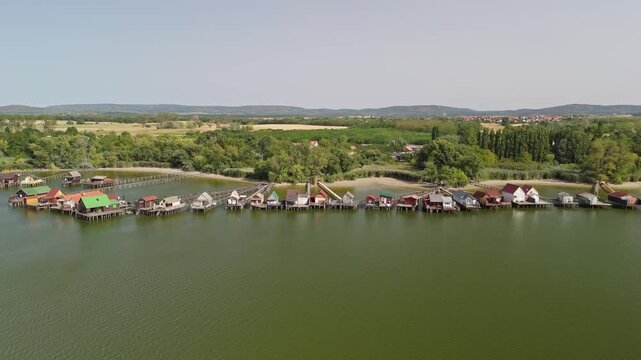 Lake Bokod in Hungary with floating houses and wooden fishing huts on a cooling reservoir, connected by piers and walkways, surrounded by forested landscape in aerial view.