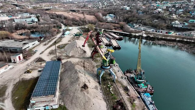 Rail-mounted portal cranes at a riverside bulk-material terminal in drone aerial view. Inland freight logistics and construction material supply