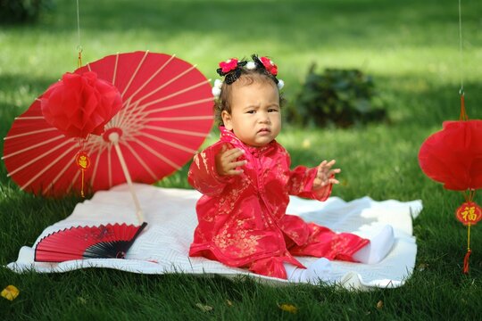 Asian baby in traditional red suit sitting on picnic blanket with fan, umbrella and paper lanterns