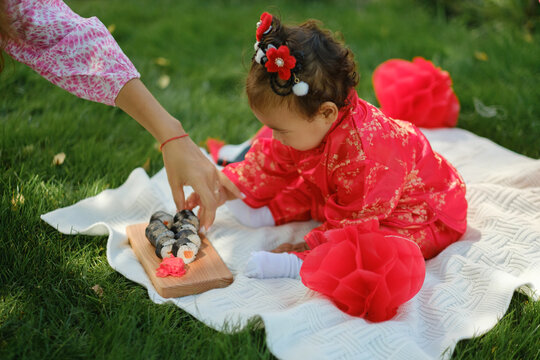 Asian baby in traditional red costume sitting on blanket with mother giving sushi rolls at picnic