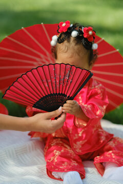 Asian baby girl in traditional red dress holding fan with red umbrella outdoors on grass