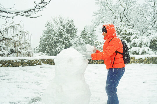 Man wearing bright outerwear forming snowman body beside snow-covered trees during active snowfall. Conveys adult winter creativity, childhood nostalgia, and seasonal outdoor joy in park setting.