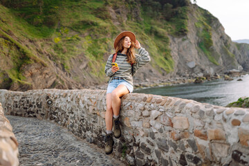 Woman wearing hat and holding phone stands on stone bridge of fortress overlooking the ocean. Traveler with backpack admires historic architecture. Concept of history, nature, and enjoyment. © maxbelchenko
