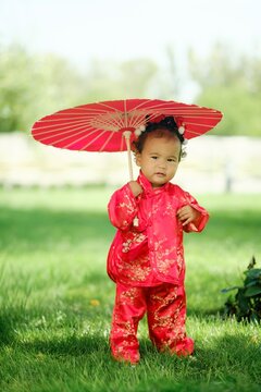 Asian baby girl in traditional red suit holding umbrella standing on green grass outdoors