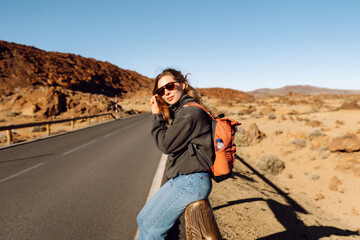 Female traveler with backpack leans against roadside barrier in remote, open landscape. Woman is resting at sunset in national park near volcano. Adventure and wilderness themes. © maxbelchenko