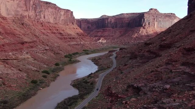 Crimson canyon aerial view of arid desert valley with muddy river, winding road beside rugged canyon walls and layered rock strata, and towering red cliffs under clear blue sky
