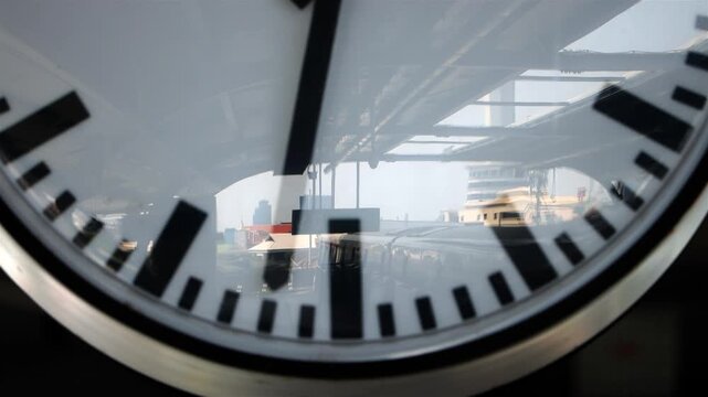 Close-up of a station platform clock with reflections of a train and urban infrastructure on the glass face