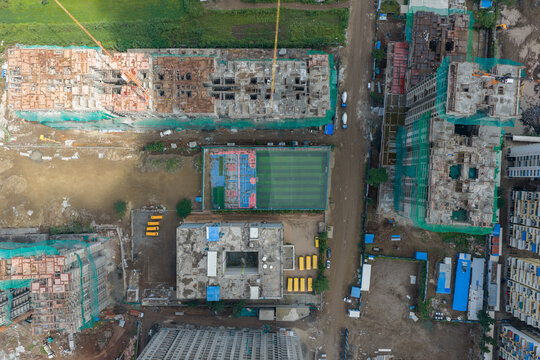 Aerial view of a construction site in Punawale featuring unfinished buildings with green safety netting, a sports field, and parked yellow school buses in Pimpri-Chinchwad, Maharashtra, India.
