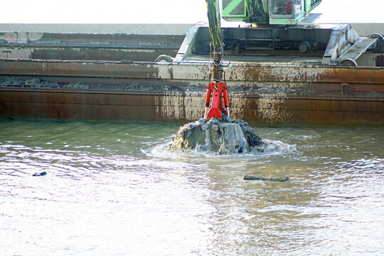 Clamshell bucket attached to hydraulic excavator emerging from river while lifting sediment beside work barge. Represents channel deepening, navigational access, and aquatic infrastructure maintenance