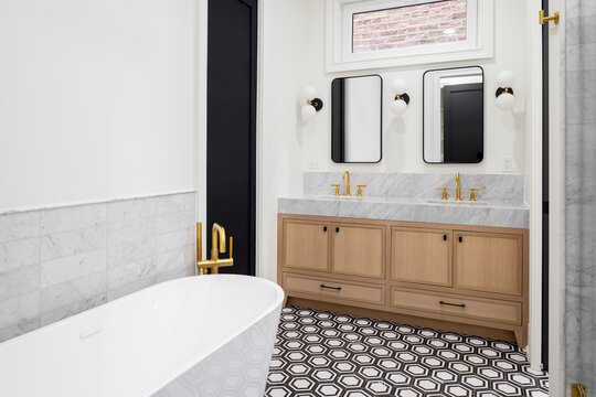 A luxury bathroom with a marble slab countertop on a natural oak cabinet, gold faucets, and a soaking tub on a white and black pattern tile floor.