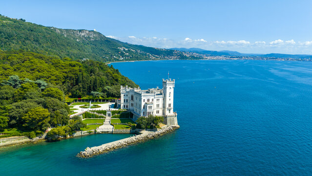 Aerial view of Miramare Castle on a rocky promontory overlooking the blue Adriatic Sea, surrounded by lush gardens and dense forest in Trieste, Friuli-Venezia Giulia, Italy.