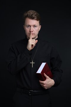 Priest in cassock with Holy Bible showing hush gesture on black background