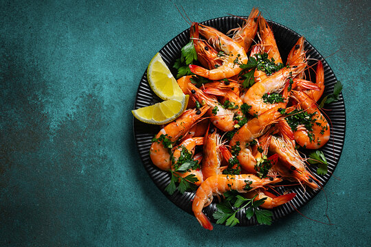 Fresh cooked shrimp with garlic, parsley and lemon on a black plate, top view, dark background.