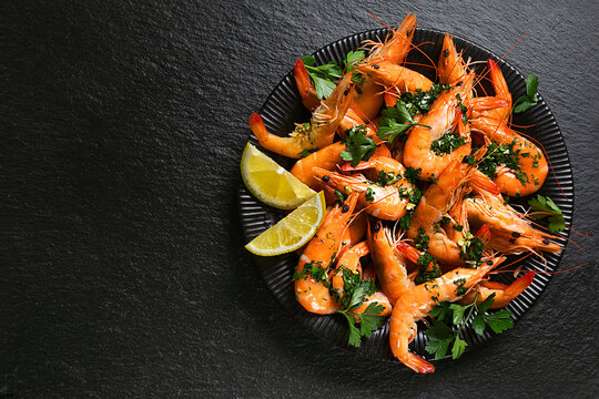 Fresh cooked shrimp with garlic, parsley and lemon on a black plate, top view, dark stone background.