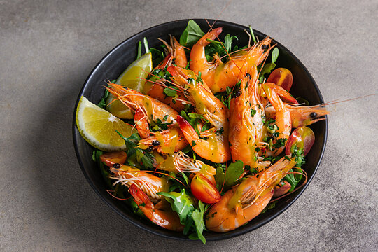 Fresh cooked shrimp with garlic, parsley and lemon on a black plate, top view, dark stone background.