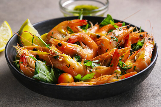 Fresh cooked shrimp with garlic, parsley and lemon on a black plate, top view, dark stone background.