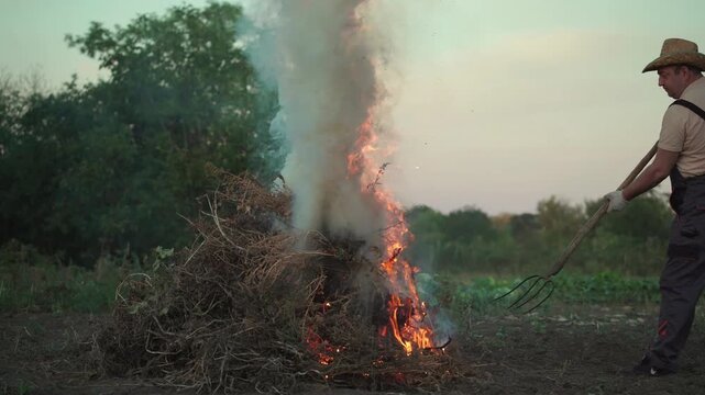 Man farmer in straw hat uses a pitchfork to manage a burning pile of dry brush and twigs in a rural field during sunset, with smoke and flames rising against a natural backdrop