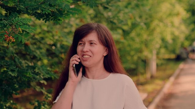 sunlight filtering through green leaves., woman with smartphone laughing softly during pleasant conversation in nature., young adult woman speaking on mobile, sunny park setting with greenery