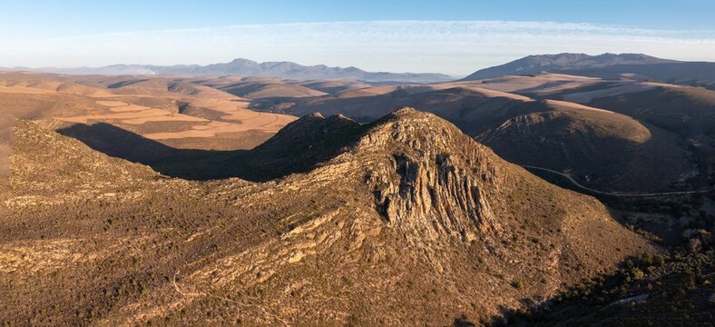 Aerial view of a rugged mountain ridge rising above vast, dry, rolling hills and arid plains under a clear sky in South Africa.