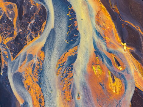Aerial view of vibrant glacial river channels with striking orange sediment and deep blue water patterns flowing through the volcanic landscape of Iceland.