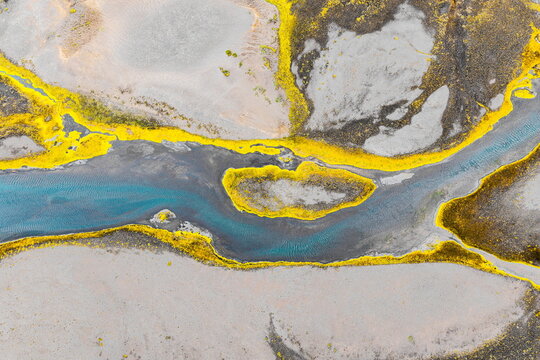 Aerial view of a glacial river with vibrant yellow mossy banks contrasting against grey volcanic sand and turquoise water in Iceland.