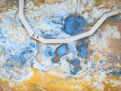 Aerial view of Myvatn geothermal area featuring vibrant blue mud pools and yellow sulfur deposits with people walking along a wooden boardwalk in Thingeyjarsveit, Iceland.