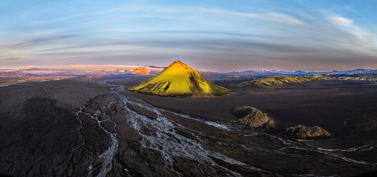 Aerial view of Maelifell, a vibrant green volcanic cone rising from dark volcanic sands with glacial river patterns and distant mountains in Skaftarhreppur, Iceland.