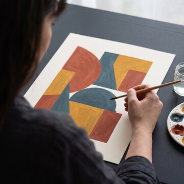 Close-up of a female artist painting abstract geometric shapes with gouache colors on paper in a creative art studio