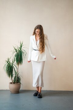 Young woman in white blazer and skirt smiling while standing beside tall potted dracaena in minimalist studio with neutral backdrop