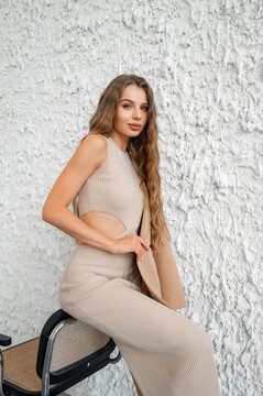 Young woman with long wavy light brown hair wearing beige ribbed knit cutout dress and draped matching coat, seated against textured white plaster wall