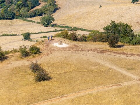Aerial view of two hikers standing near a trig point on the dry grassy slopes of Cleeve Hill with rolling landscape and shrubs in the background Cheltenham, England, United Kingdom.