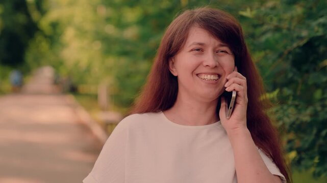 relaxed weekend city park., woman enjoys phone conversation while walking along tree-lined path today., smiling woman in white shirt chatting on phone outside today., brunette woman calling friend