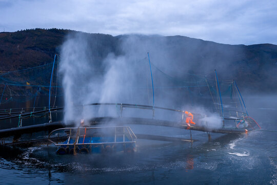 Aerial view of a worker in orange protective gear using high-pressure water to clean the underside of an aquaculture fish farm ring in the fjord, Liabygda, More og Romsdal, Norway.