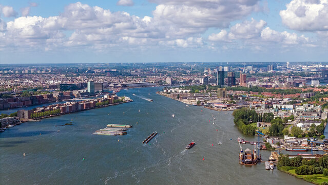 Aerial view of the IJ river waterfront with cargo ships, industrial buildings, and urban infrastructure under a blue sky in Amsterdam, North Holland, Netherlands.