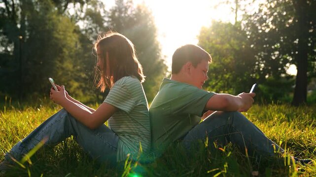 Kids leaning back to back with phones, Warm evening light in park, Nature versus screen contrast, Peaceful meadow tech moment, Online chats in fresh air, Summer leisure digital style, Youth focused on