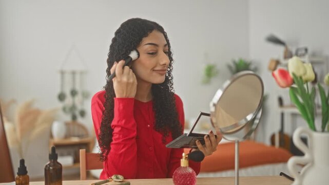 Young woman applies makeup brush to cheek while holding compact palette at a vanity mirror in bedroom; self care calm.