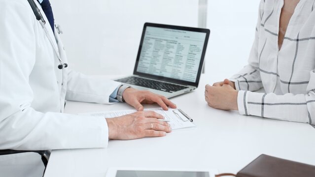 Doctor in a white coat with a stethoscope is consulting a patient across a table. Taking notes on a clipboard. And reviewing medical data on a laptop