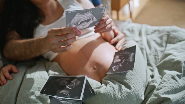 Closeup couple holding sonogram photos over pregnant belly. Ultrasound on bed.