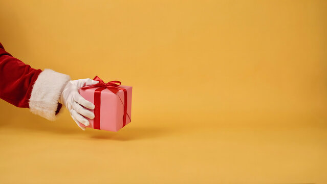 Santa claus hand wearing a white glove holding a small pink gift box with a red ribbon bow, isolated on a solid yellow background space