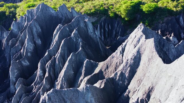 Jagged grey rock formations and steep eroded ridges are illuminated by bright sunlight beneath a strip of lush green vegetation.