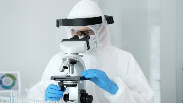 Scientist in full protective suit using microscope for biological research, developing vaccine, examining samples for infection, analyzing pandemic virus
